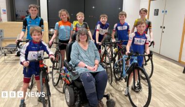 Six children in a assorted sports lycra stand holding bikes, in front of them are two boys also in cycling lycra holding bikes either side of a lady with round glasses and grey hair. She is in a wheelchair and is wearing a teal top and navy trousers, her right knee has a knee brace on it.