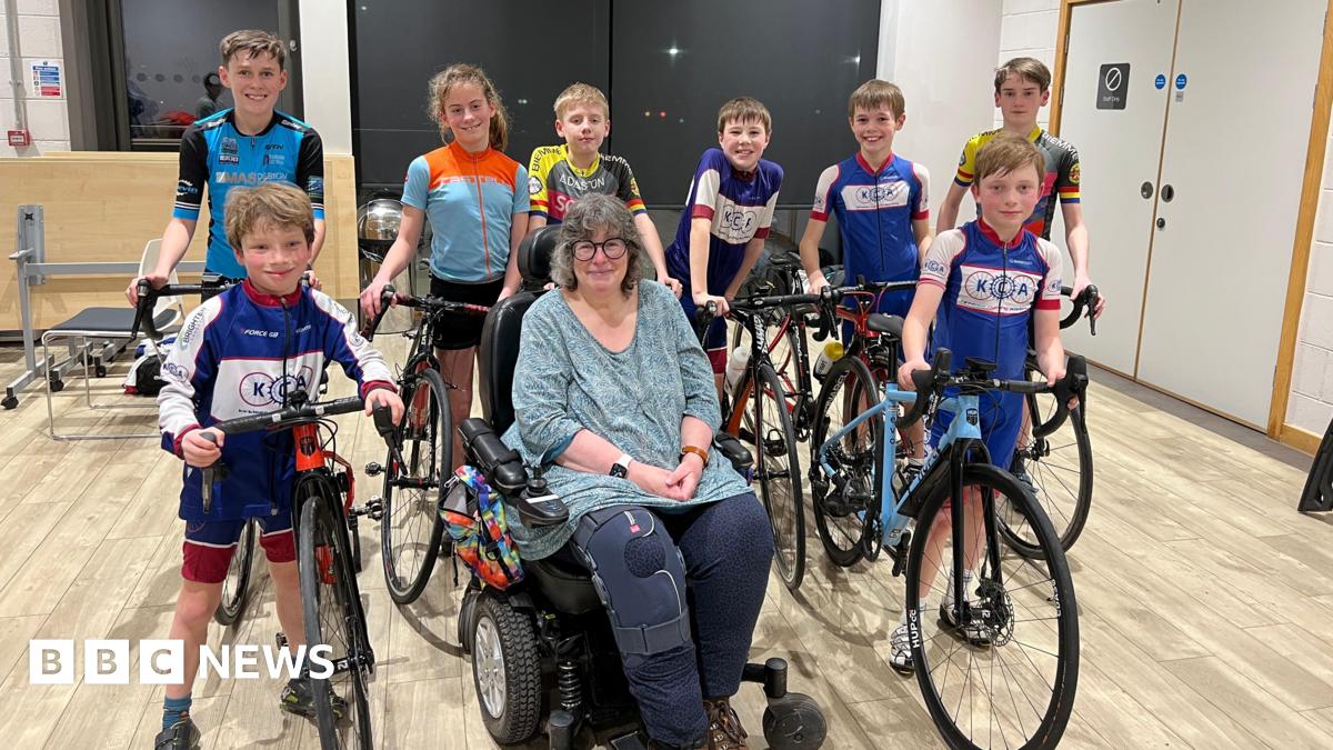 Six children in a assorted sports lycra stand holding bikes, in front of them are two boys also in cycling lycra holding bikes either side of a lady with round glasses and grey hair. She is in a wheelchair and is wearing a teal top and navy trousers, her right knee has a knee brace on it.