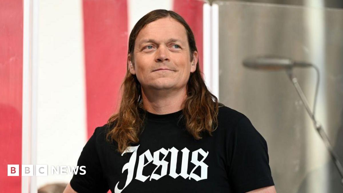 Brad Arnold of the rock band 3 Doors Down smiles . He has long hair and black T-shirt with 'Jesus' written across the front.