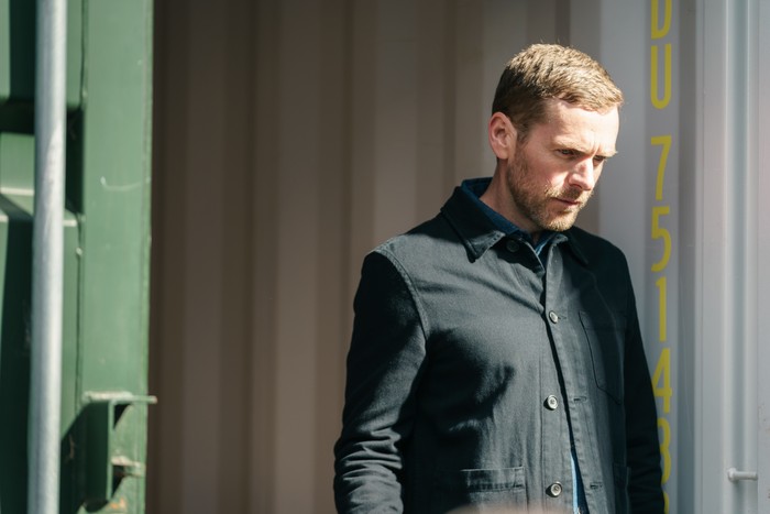 Shaun Evans as John Hughes in Betrayal, wearing a black shirt and stood by a wall.