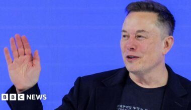 Elon Musk in a black t-shirt and black blazer waving at the audience during a conversation at this year's World Economic Forum in Davos.