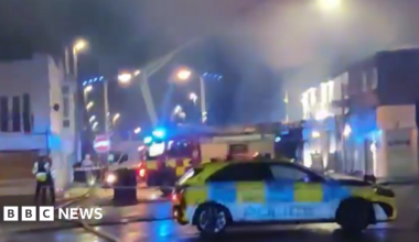 A police car is parked outside the corodned area with fire engine in background and apparatus hosing down the smoks over shops and premises at night.