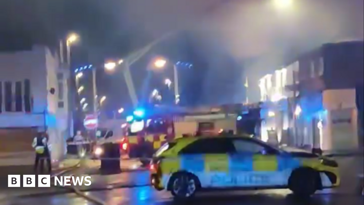 A police car is parked outside the corodned area with fire engine in background and apparatus hosing down the smoks over shops and premises at night.