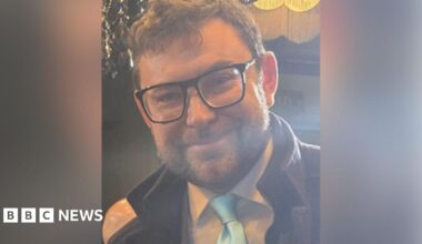 Adam Mitula with brown hair and brown beard wearing black glasses and a white shirt, pale blue tie and dark brown coat. He is smiling.