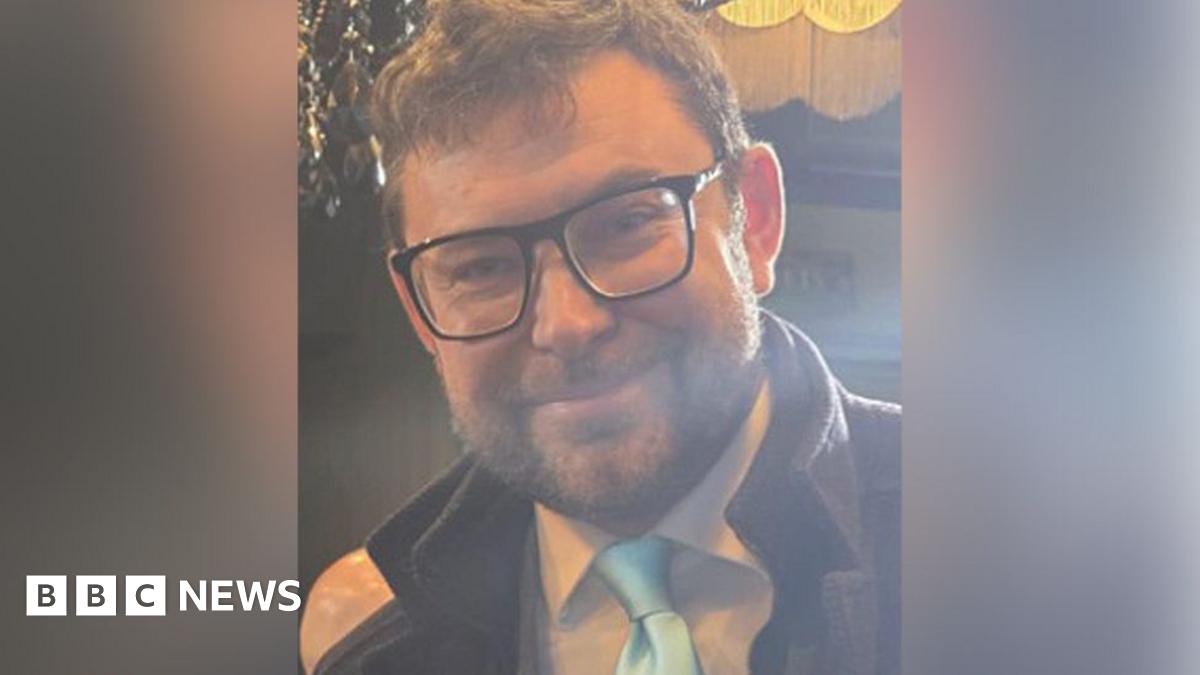 Adam Mitula with brown hair and brown beard wearing black glasses and a white shirt, pale blue tie and dark brown coat. He is smiling.