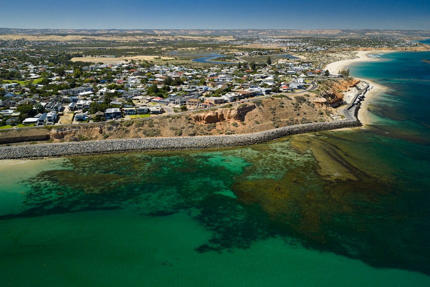 Aerial shot showing houses next to a small cliff along a stretch of beach. Dark shadows indicating a large reef in the water