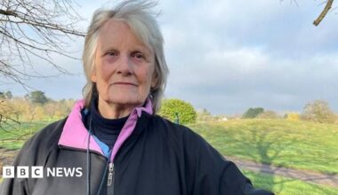 The golf course is on the other side of a wooden fence and there are golfers dressed in black and white playing on the pitch. Their trolleys are nearby. The golf course has trees around it and along the hill in the background.
