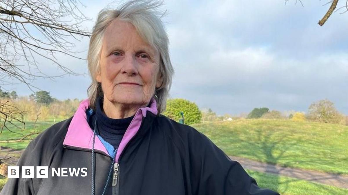 The golf course is on the other side of a wooden fence and there are golfers dressed in black and white playing on the pitch. Their trolleys are nearby. The golf course has trees around it and along the hill in the background.
