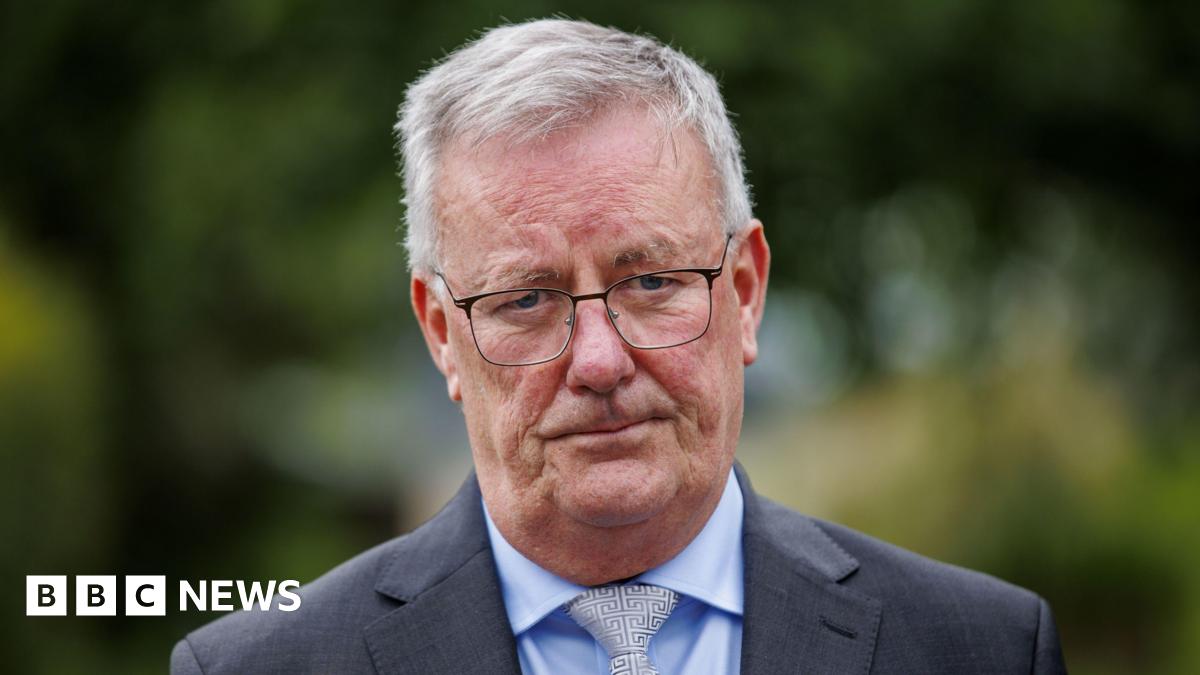 Mike Nesbitt is wearing a blue shirt, white tie and grey blazer. He has short grey hair and glasses on his face. The background is blurry.