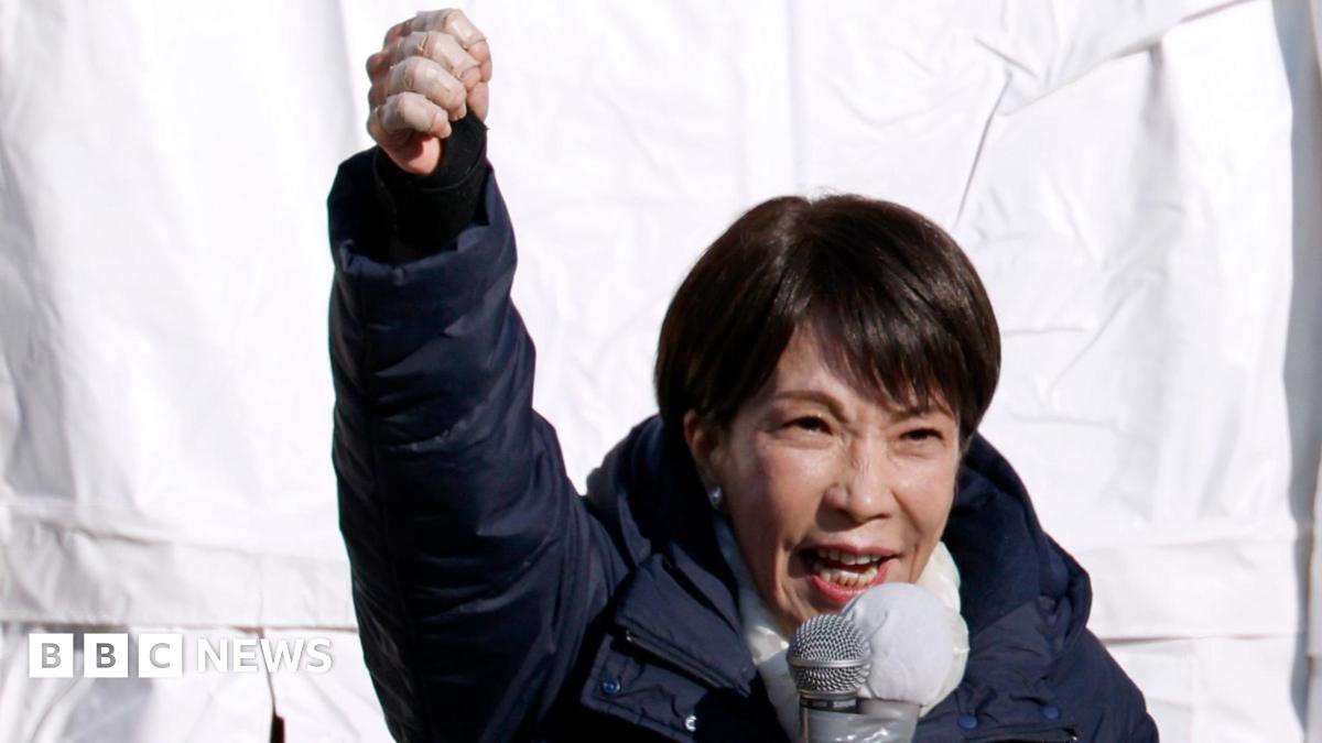 Sanae Takaichi speaks into a microphone with her fist raised. She's wearing a dark blue puffer coat