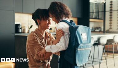 A parent kneels in a modern kitchen, holding and comforting a school‑aged child who is wearing a uniform and a large backpack, as they share a close and supportive moment.