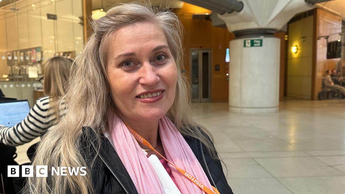 Katalin Kaszas smiling while seated indoors, wearing a pink scarf and black jacket.