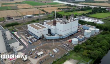 Aerial view of the power station at Keadby in North Lincolnshire. There is a large central white building with smaller buildings around the perimeter of the site. There are trees and farmland in the background.