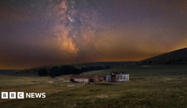 An abandoned barn in a field lit by the night sky.