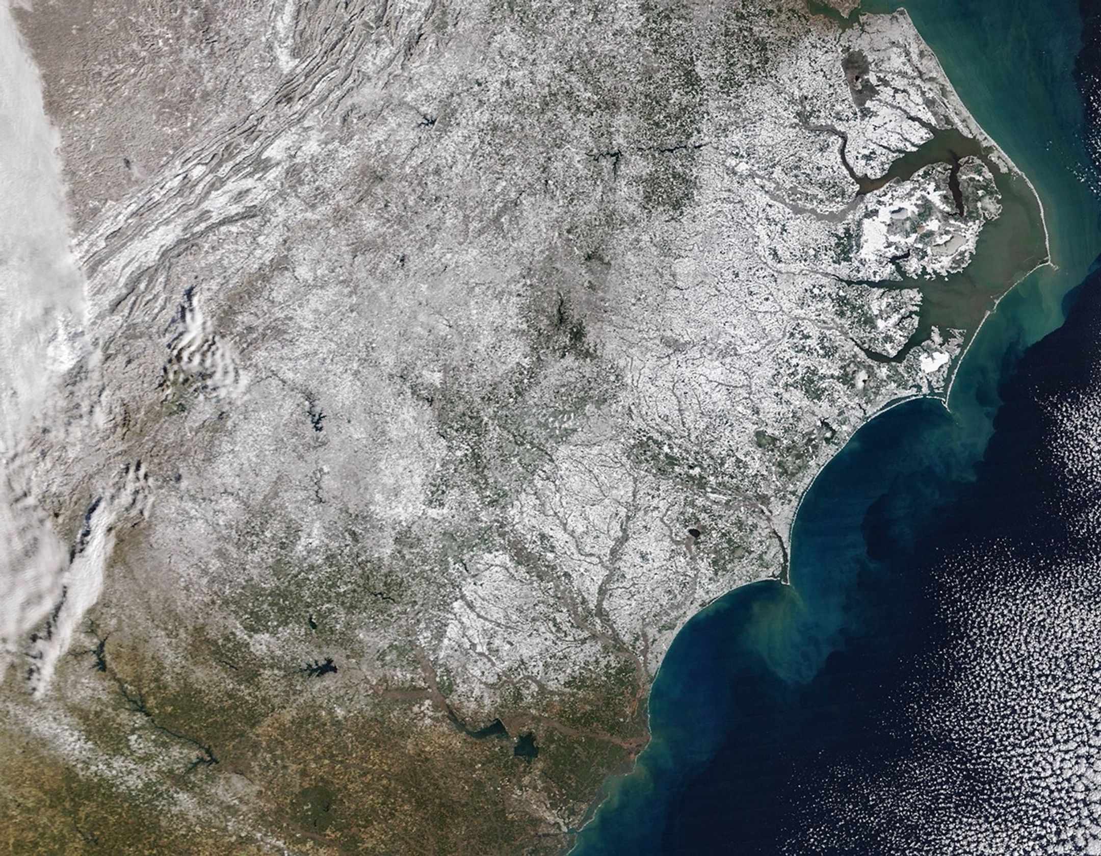A satellite image centered on North Carolina shows white snow covering most of the state, with additional snowy patches in neighboring states. Some forested and urban areas appear gray. To the right, a strip of barrier islands appears white against the green and blue Atlantic Ocean.