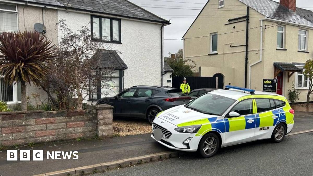 A police car, partially parked on the pavement, outside a white-painted, semi-detached house with black trim on the windows. There is a grey sports car parked on the gravel drive in front of the house, where a police officer in a hi-vis yellow jacket stands, with his arms crossed