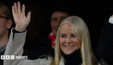 A woman with blonde hair smiles and waves