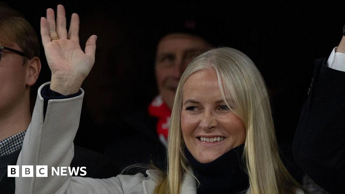 A woman with blonde hair smiles and waves