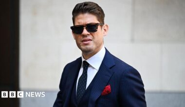 A man with short brown hair wears dark sunglasses and a blue pinstriped suit with a red pocket square looks without smiling towards the camera.