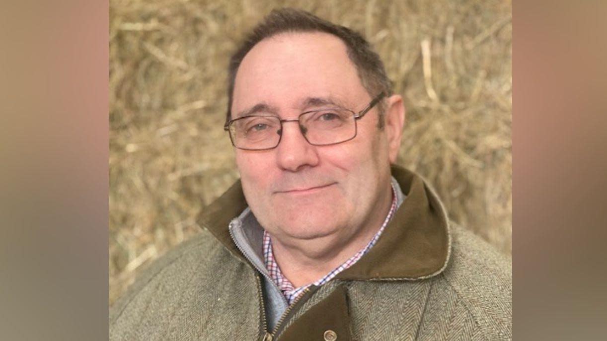 Farmer Steve Glover looks at the camera. He is wearing a green and brown tweed jacket and in the background there is a large pile of hay.