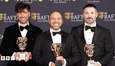 Three men stand on the 2026 BAFTA red carpet in black tuxedos. Each of the three hold a BAFTA award in the shape of a gold theatrical mask.