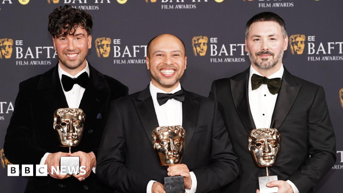 Three men stand on the 2026 BAFTA red carpet in black tuxedos. Each of the three hold a BAFTA award in the shape of a gold theatrical mask.