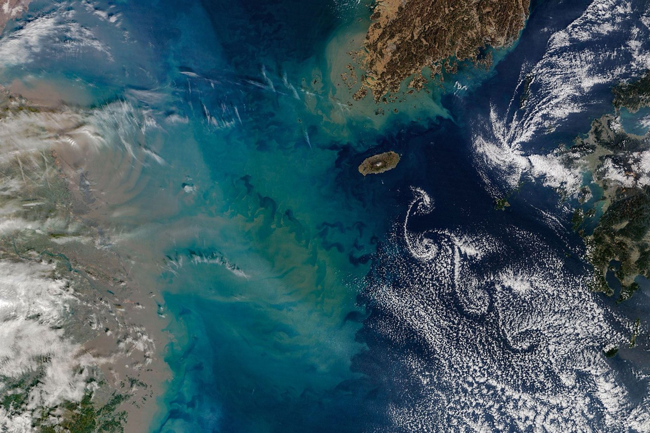 A series of spiraling clouds extends southeast from an oval-shaped island in the Korea Strait. To the west, a large sediment plume fans out from the coast of China and forms tan, teal, and blue swirls in the water.