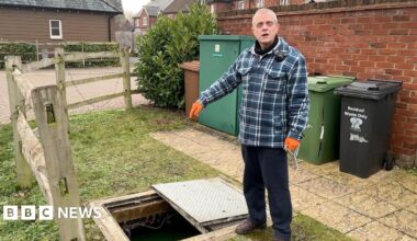 Stuart Tyrrell is wearing a blue checked overshirt and orange work gloves. He's pointing into a hole created where he just removed a manhole cover. It is a residential area and behind him are brown, green and black wheelie bins.