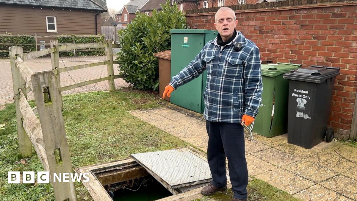 Stuart Tyrrell is wearing a blue checked overshirt and orange work gloves. He's pointing into a hole created where he just removed a manhole cover. It is a residential area and behind him are brown, green and black wheelie bins.