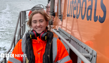 A smiling Hatty McKay stands on the deck of an orange lifeboat. She is wearing an orange hi-viz jacket and ear defenders around her neck.