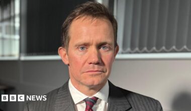 A head and shoulders photo of Mark Teeton, a middle-aged white man with brown hair and piercing light blue eyes, looking into the camera. He wearing a dark grey suit, a white shirt and a red and blue striped tie.