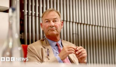Rupert Lowe looking thoughtful. He is wearing a cream suit jacket over a light blue shirt and a red patterned tie.