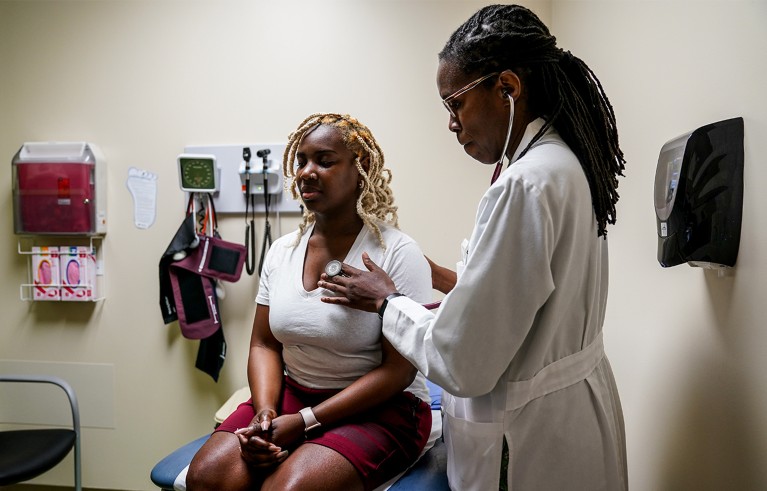 A patient sits on a chair while a doctor uses a stethoscope to listen to her chest