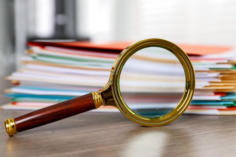 A close up view of a magnifying glass and a pile of many folders containing paper documents on a wooden desk.