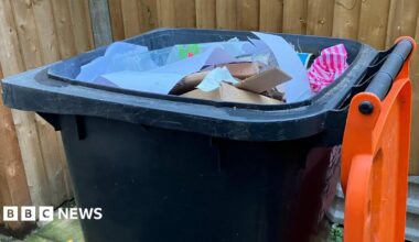 Dark grey wheelie bin with the orange lid opened, and paper and cardboard visible inside. The bin is in front of a wooden fence.