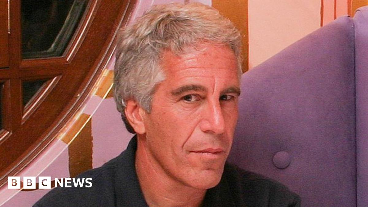 Jeffrey Epstein looking at the camera with his face slightly turned to his left. He is wearing a blue top and is sat on what looks like a purple sofa, with a window partially visible to his right.