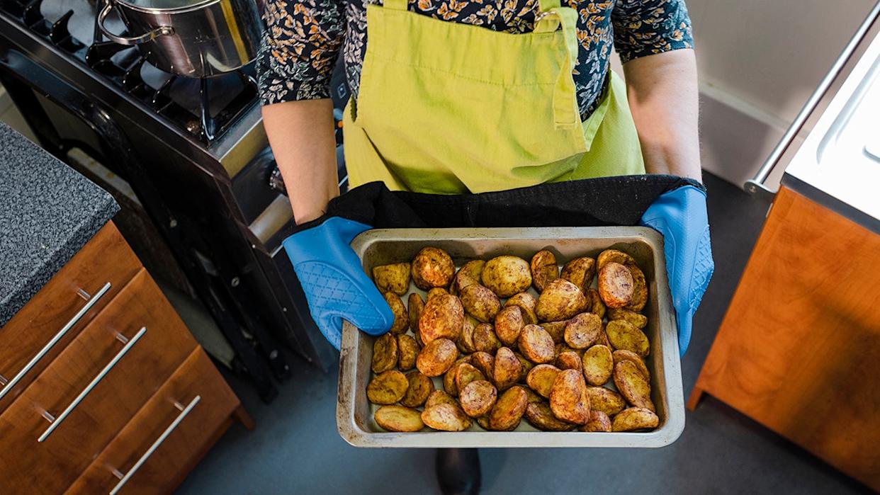 Woman holding tray of roasted potatoes, seen using blue oven mitts in kitchen