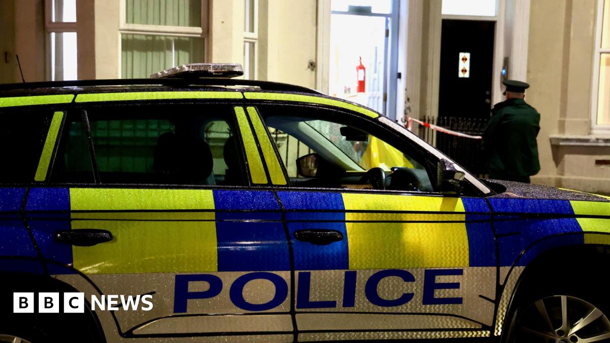 A police car outside a cream house. The door is open and an officer standing outside. The car is green white and blue.