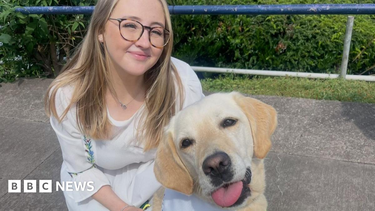 Megan Stephenson is crouching down with her assistance dog. She is smiling and has long blonde hair and is wearing glasses. She is wearing a white dress. Her dog is in the foreground of the photo.