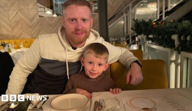 Michael Woods, 36, who has ginger hair and a short ginger beard, sits at a table in a restaurant with his arm around his son, six, who has short blonde hair.