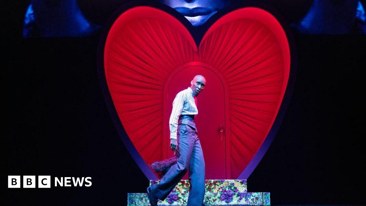 Cynthia Erivo stands in front of a heart on stage in a suit while a visual projection of her face is above it.