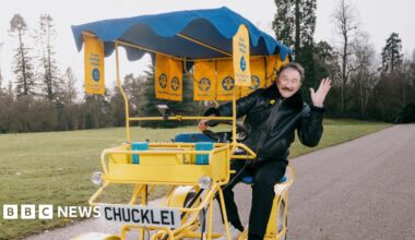 A man in a black jacket and jeans rides a two-seater pedal car in yellow and blue. The number plate on the front reads Chuckle1