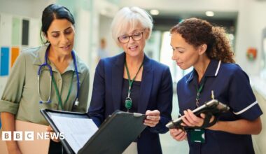 A woman middle-aged woman wearing a shirt with dark hair and a stethoscope around her neck, an older woman in a blazer holding a file, and a younger woman in a nurse's outfit holding a tablet computer, all stand next to each other in a hospital ward
