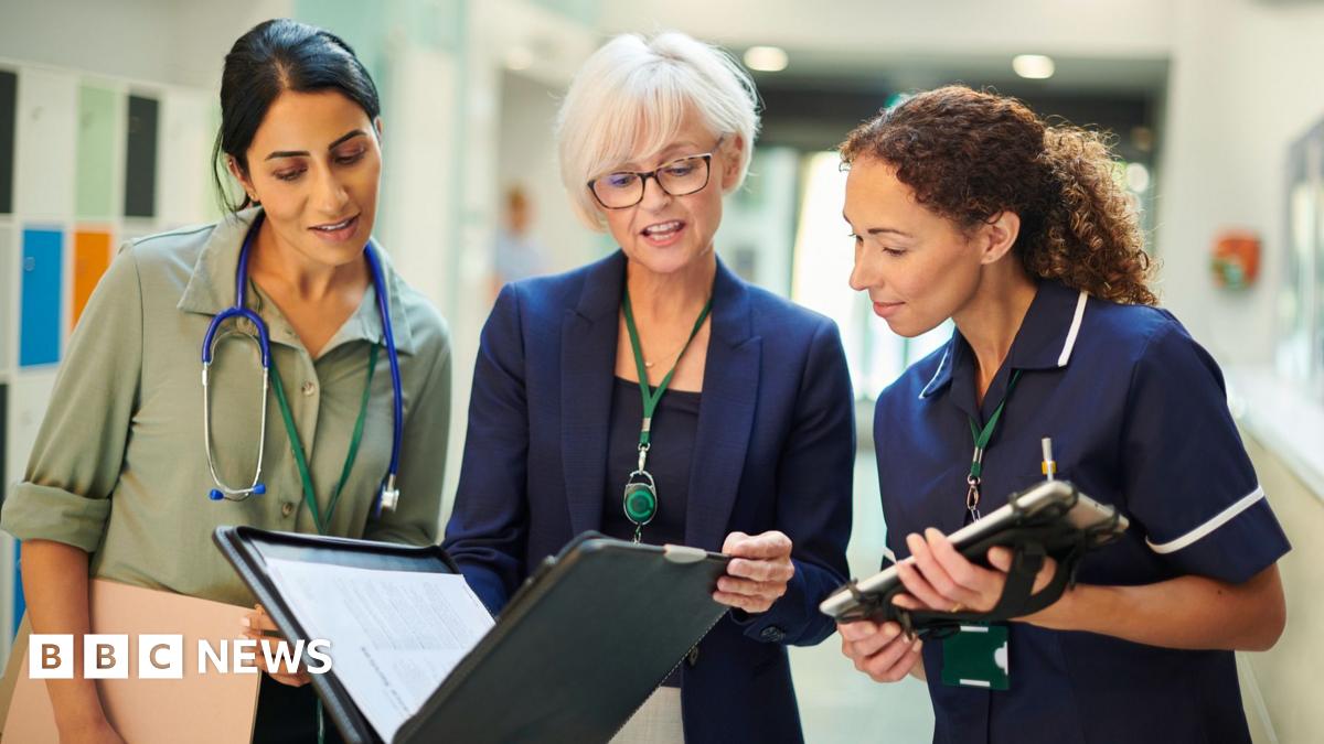 A woman middle-aged woman wearing a shirt with dark hair and a stethoscope around her neck, an older woman in a blazer holding a file, and a younger woman in a nurse's outfit holding a tablet computer, all stand next to each other in a hospital ward