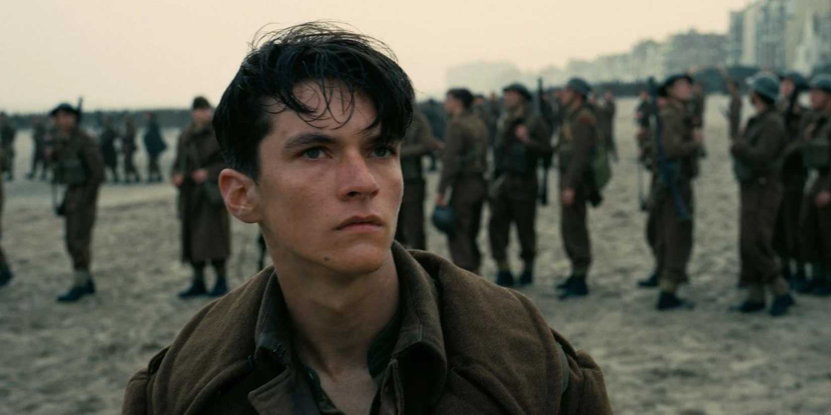 Fionn Whitehead as Tommy, standing solemnly on the beach with other soldiers in the background in Dunkirk.