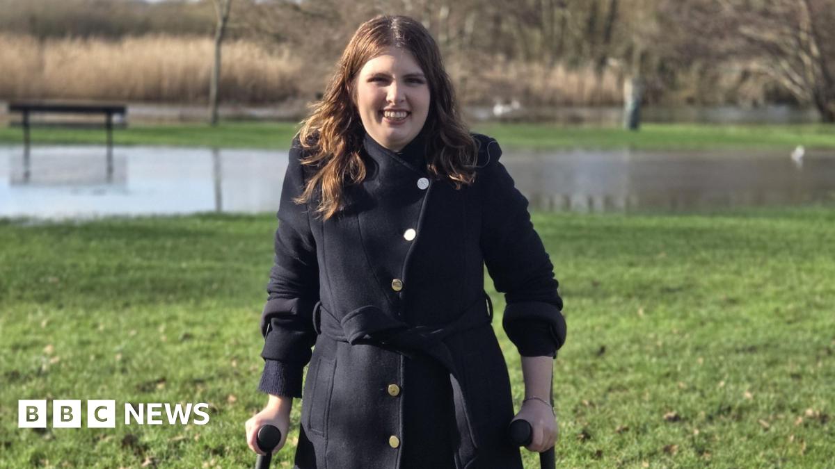 A woman with mid-length brown hair, standing outside on grass in a black jacket. She is smiling widely and her hands clasp crutches. Behind her is a blurred patch of water and some trees.