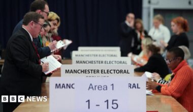Votes are counted for the Gorton and Denton by-election at the Manchester Central Convention Complex. A row of people standing and wearing party rosettes stands to the left of a series of tables while vote counters sit on the other side.