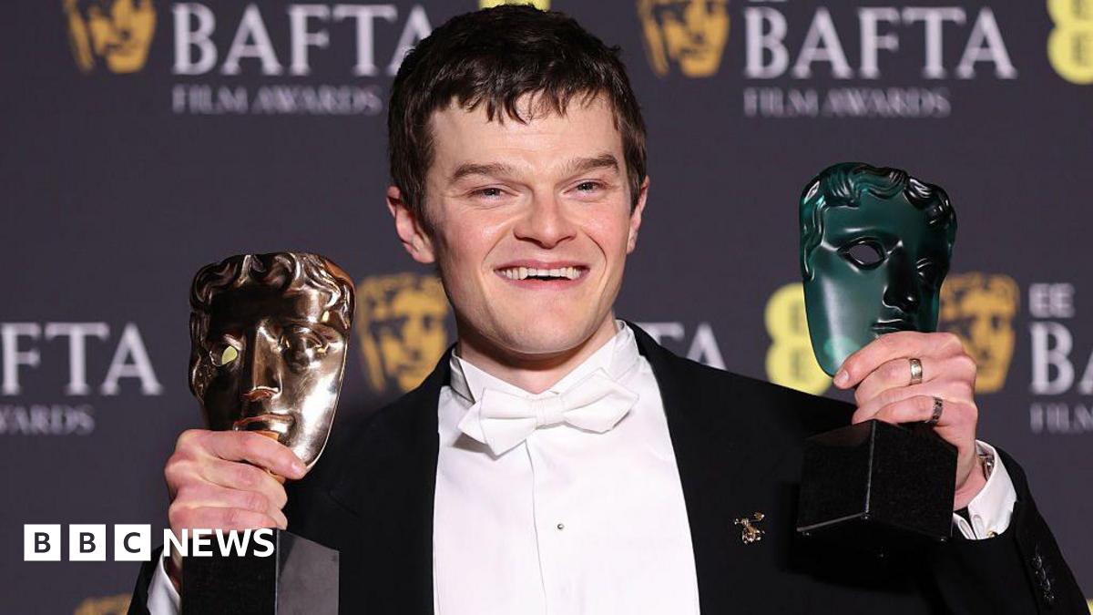 Head and shoulders shot of Robert Aramayo celebrating at the Baftas. He is wearing a white shirt and bow tie and a black jacket. He is holding up his awards and smiling.