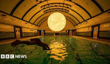 A man dives into a dimly lit swimming pool. Above the pool, suspending from an arched ceiling is a large glowing sun.