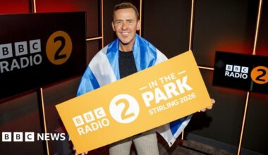 Scott Mills, wearing a Saltire flag and holding a Radio 2 In the Park Stirling 2026, in a radio studio